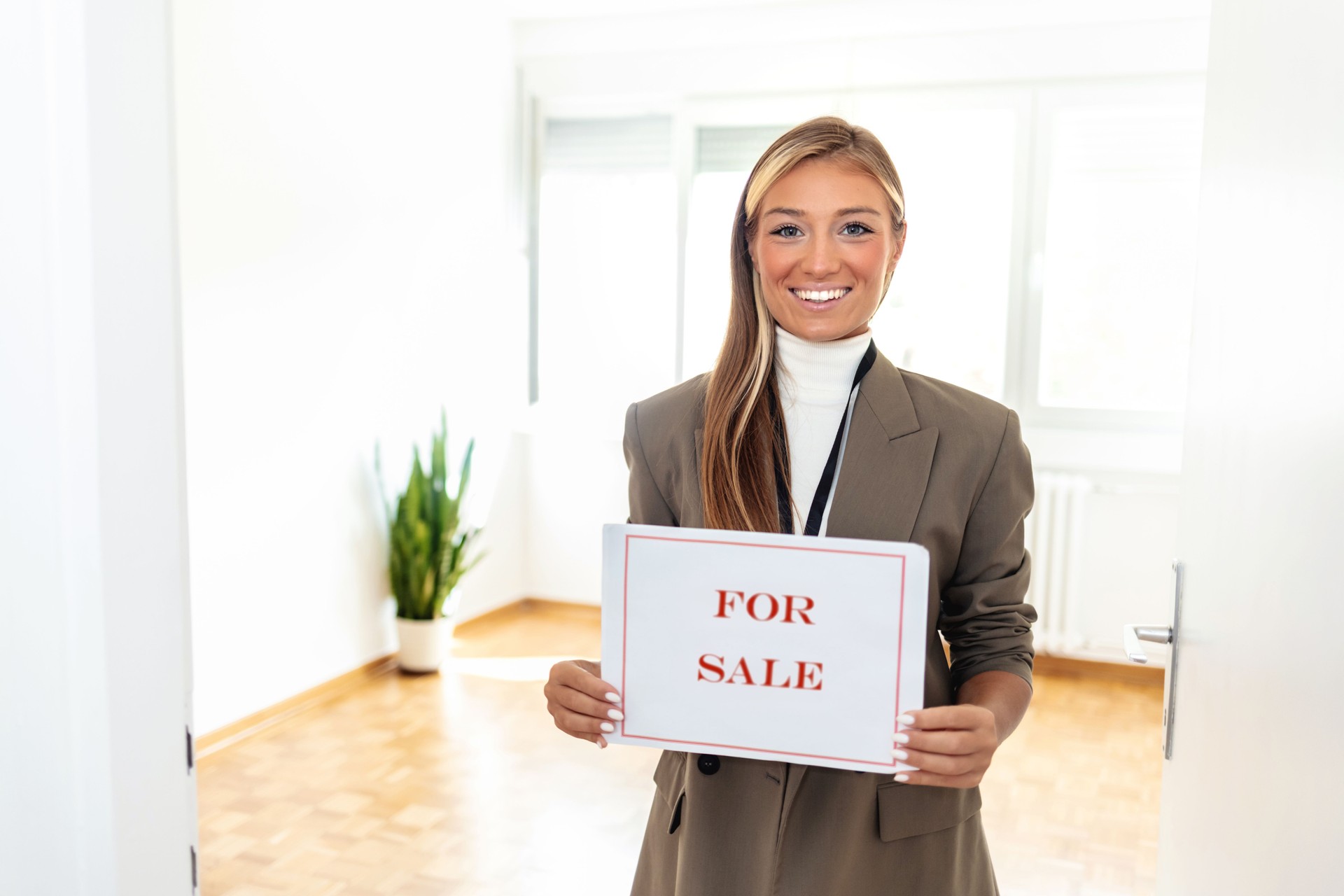 Real estate agent holding a FOR SALE sign in front of a house. Happy Real Estate Agent woman showing home for sale sign.